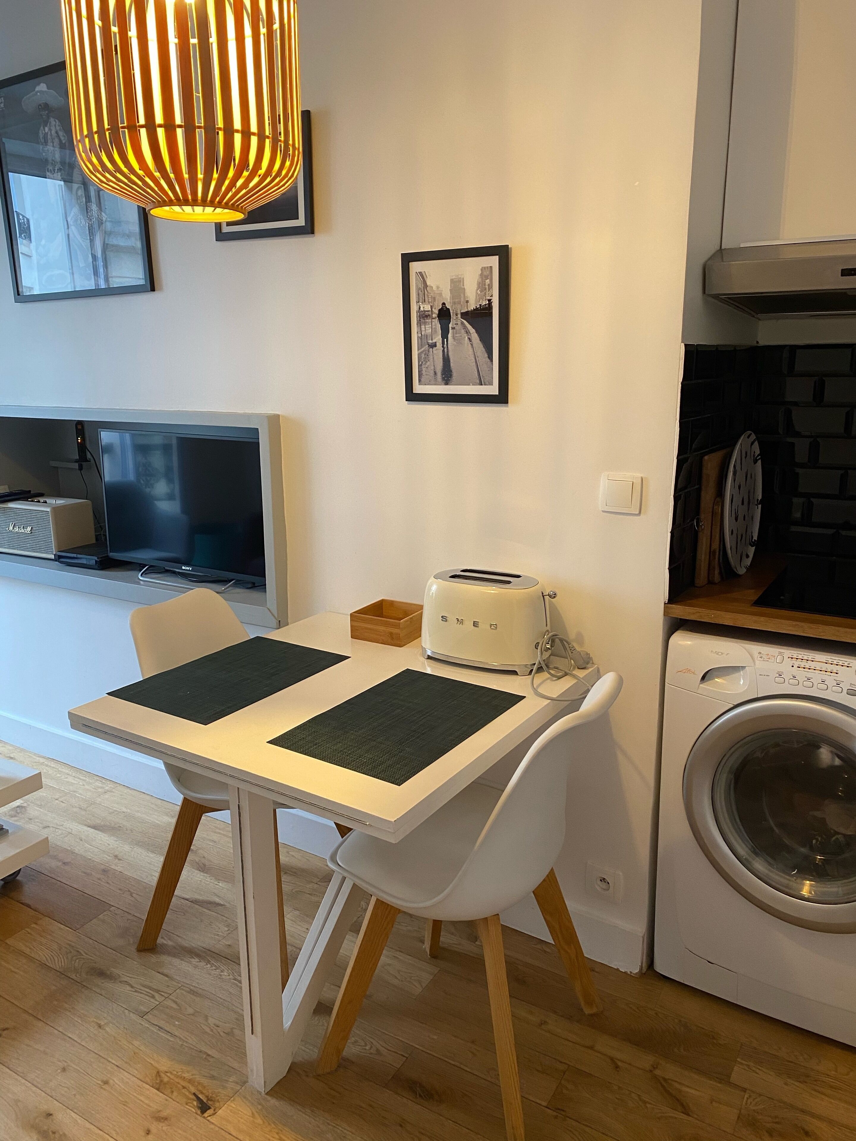 A modern kitchen with a dining table, chairs, a toaster, a washing machine, and a hanging light fixture.