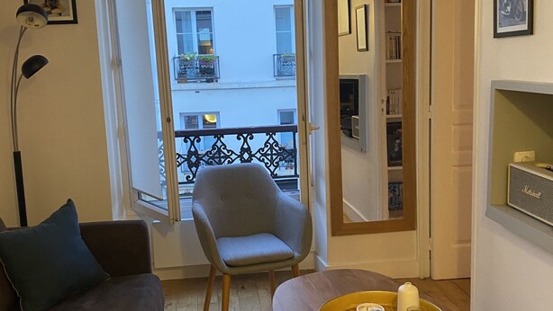 A living room with a sofa, armchair, and a small table with a tray. A window with a view of a balcony is visible. - Pretty cocoon canal St Martin (Paris)