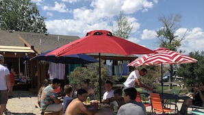 A group of people sitting at a wooden table under a red and white striped umbrella, with a wooden structure and trees in the background. - The Cow Lodge (Clark)