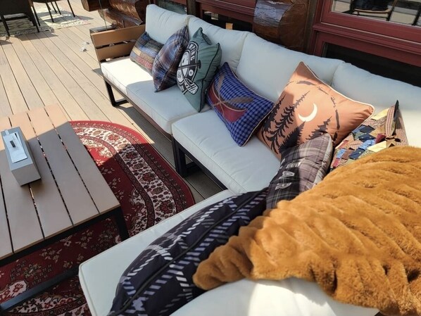 A sunlit outdoor seating area with a white sofa, patterned cushions, and a wooden table. - Shangrilog - Cabin with Mountain View & Lake Front Property (Troy)