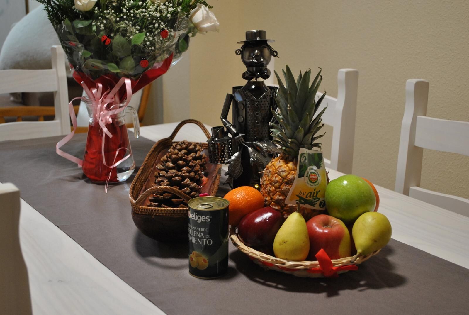 Ein Tisch mit einem Blumenstrauß, einer Ananas, einem Korb mit Äpfeln und Orangen, einer Bierdose und einer dekorativen Metallfigur.