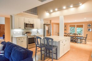 A modern kitchen with a central island, stainless steel appliances, and a dining area with a wooden table and chairs. - Short Walk to Private Beach! (West Yarmouth)