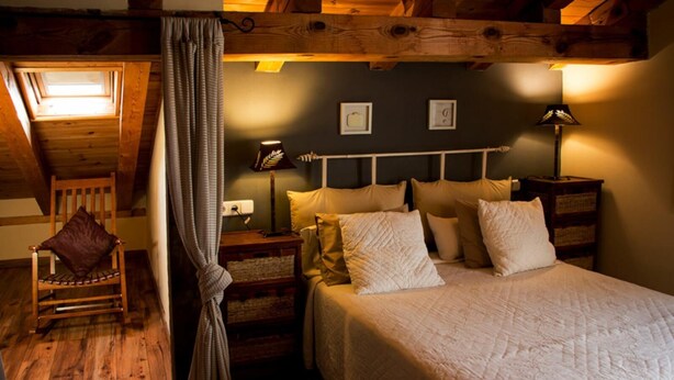 Un dormitorio acogedor con techo de madera, una cama con varias almohadas, una mesita de noche con lámpara y un sillón con cojín.