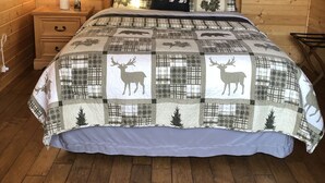Una camera da letto in legno accogliente con un letto ricoperto da una coperta con cervi e orsi, un comodino con una lampada e una finestra con tende.