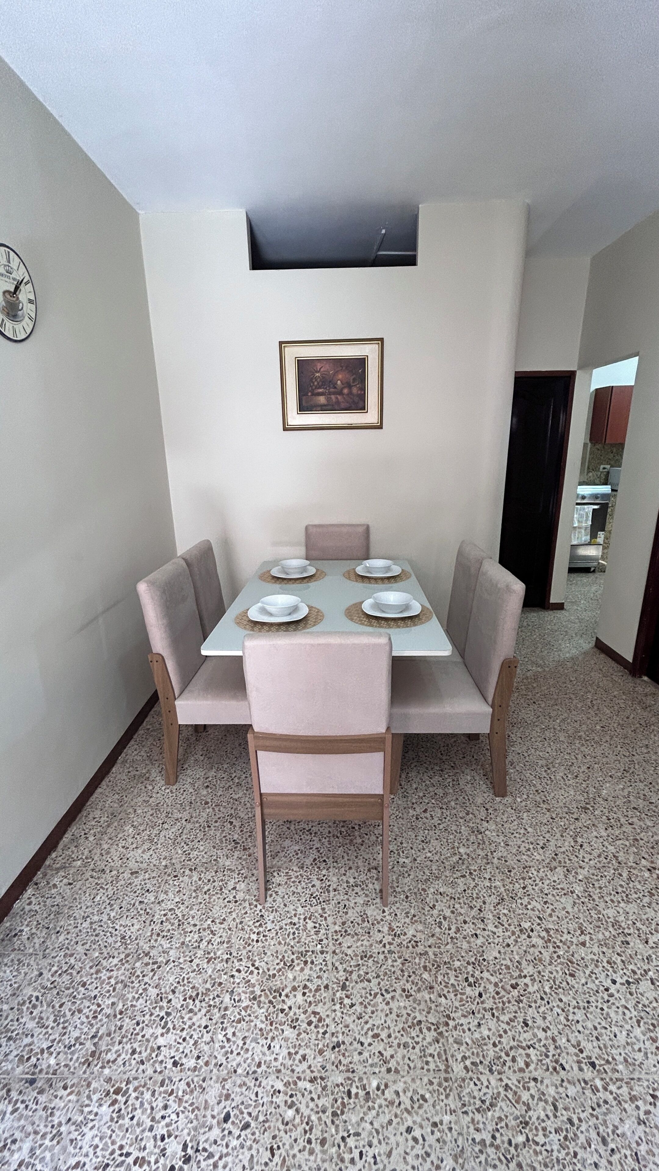 A dining area with a table set for four, chairs, a clock, and a framed picture on the wall.