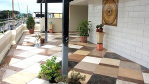 Un espacio al aire libre cubierto, con suelo de baldosas, plantas en macetas y una obra de arte colgada en la pared.