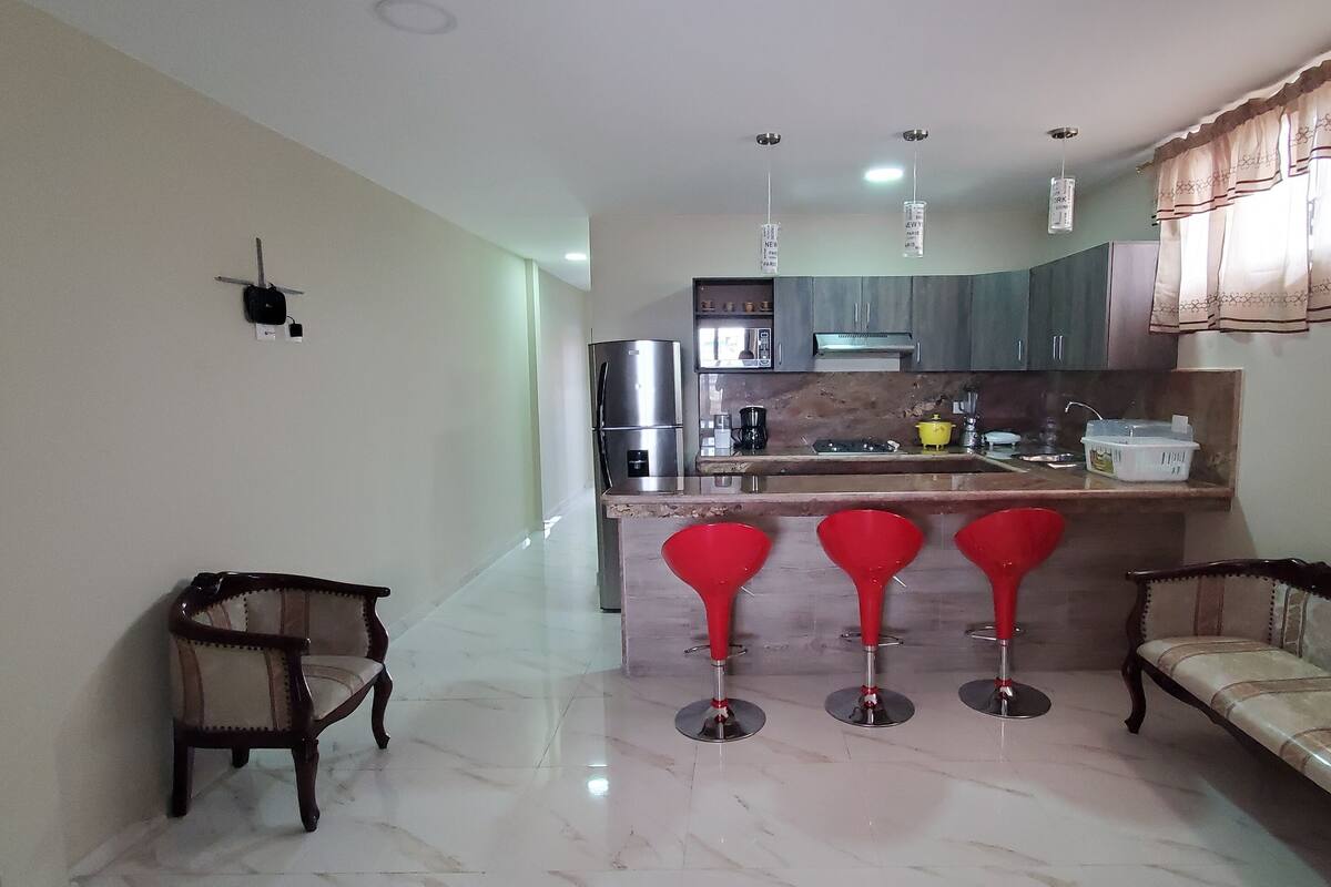 Una cocina moderna con refrigerador, estufa y taburetes de bar. Hay dos sillas de madera y un banco.