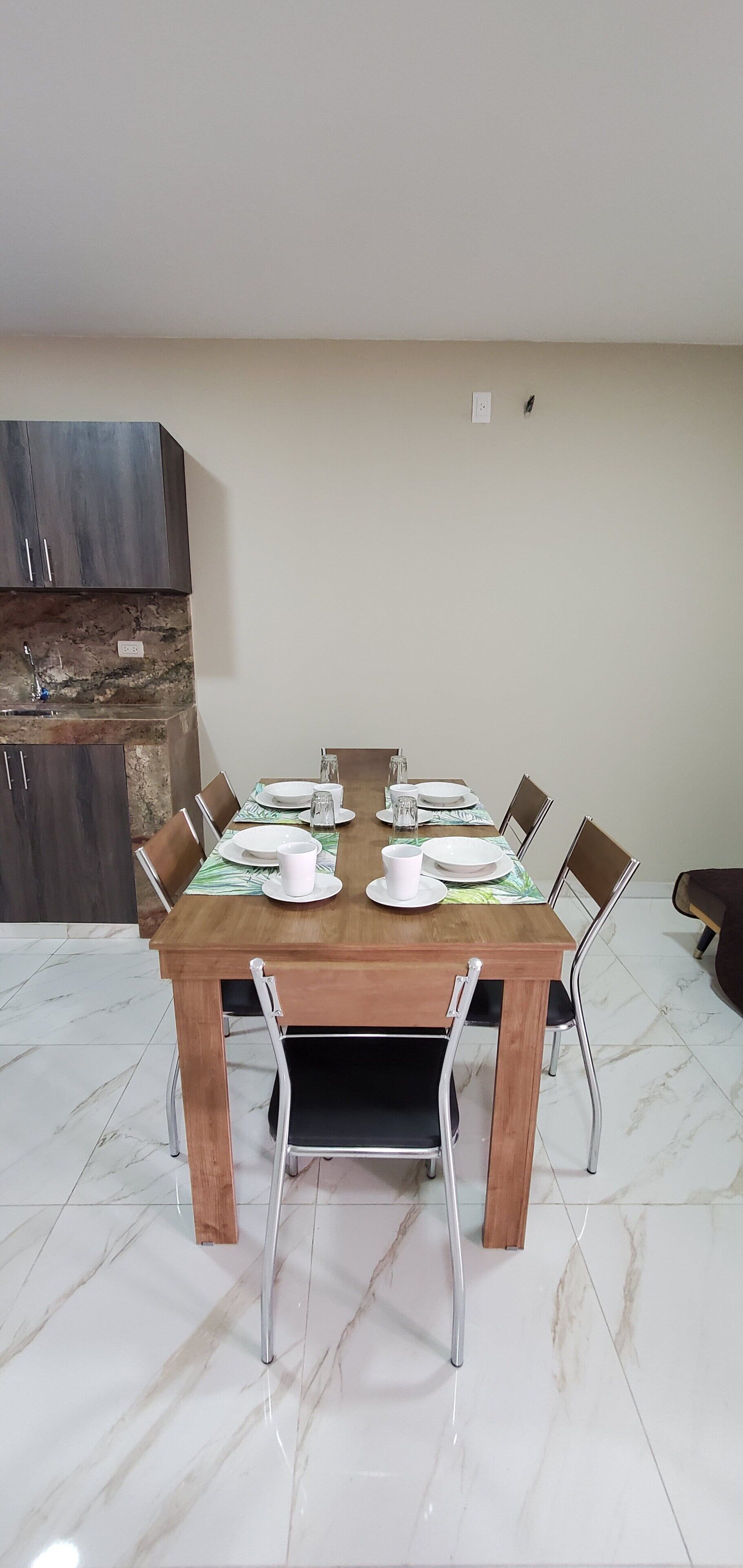 Un comedor con una mesa de madera dispuesta para cuatro personas, rodeada de sillas metálicas, en una habitación con suelo de mármol y armarios de madera oscura.