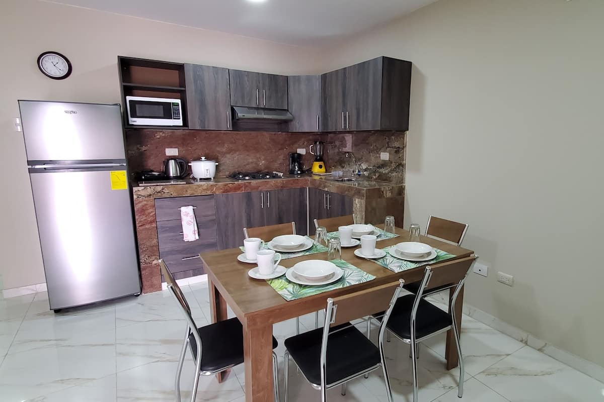 Cocina compacta equipada con refrigerador, microondas y estufa a gas. Hay una mesa de comedor con platos, tazas y un mantel individual.