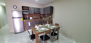 A compact kitchen with a refrigerator, microwave, stove, and sink. There is a dining table with chairs and a clock on the wall. - NCG Comegua 1 (Guayaquil)