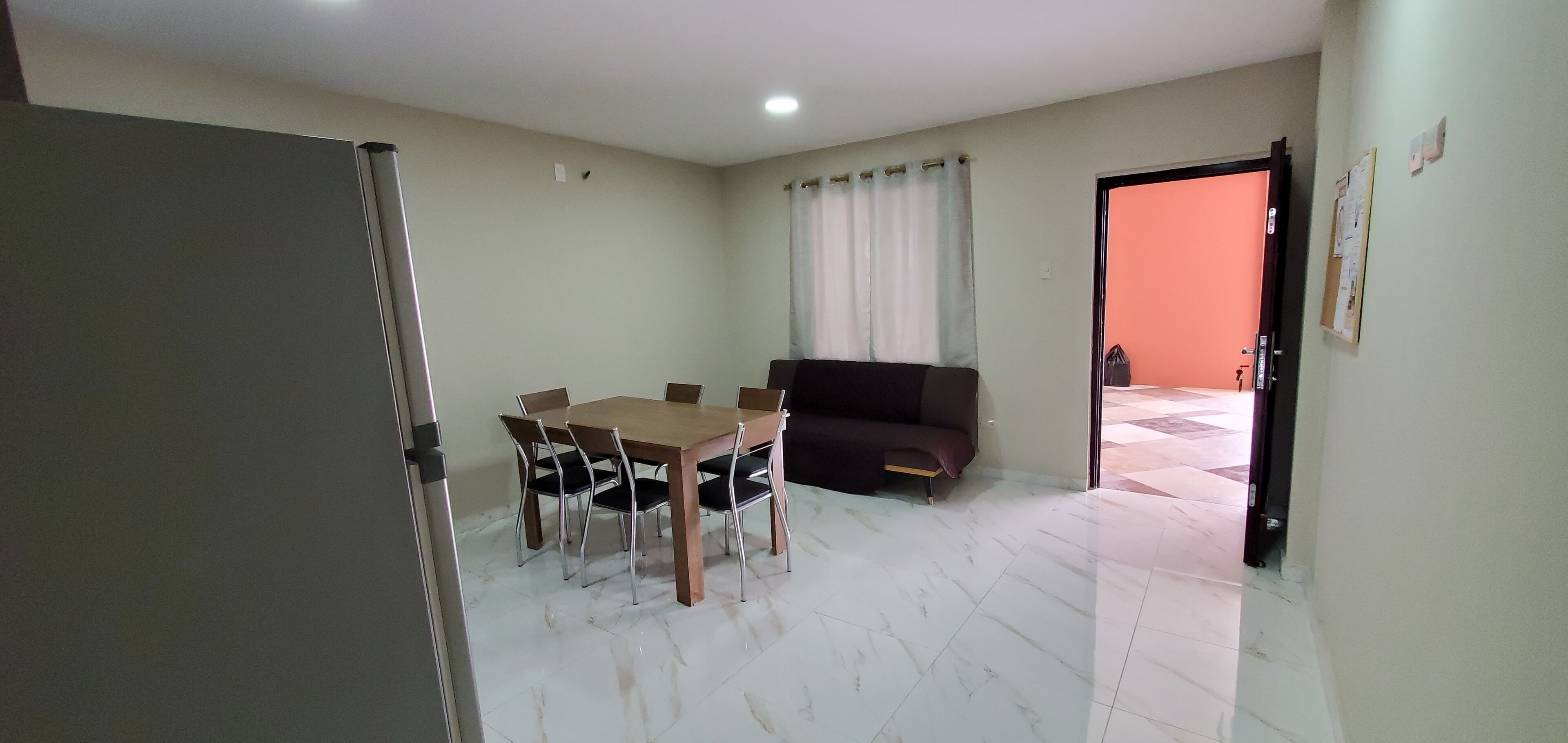 Una habitació amb una taula de dinar i cadires, un sofà, una porta que dona a una altra habitació i un tabló d'anuncis a la paret.