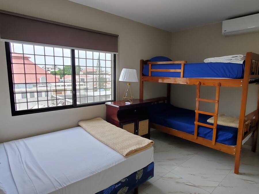 A dormitory room with bunk beds, a desk, a chair, a lamp, and a window with a view of buildings outside.