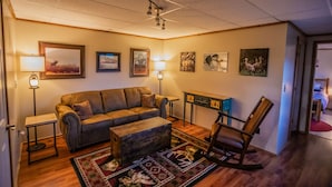 A cozy living room with a leather sofa, a wooden coffee table, and a rocking chair. There are framed pictures on the walls and a lamp on a side table. - Otter Stop Inn! Ocean and mountain views from every window! (Homer)
