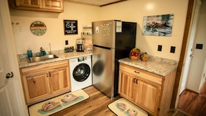 A kitchen with wooden cabinets, a stainless steel refrigerator, a washing machine, and a coffee maker. - Otter Stop Inn! Ocean and mountain views from every window! (Homer)