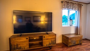 A wooden entertainment center with a flat-screen TV, a small cabinet, and a window with a view of trees and a lantern. - Otter Stop Inn! Ocean and mountain views from every window! (Homer)
