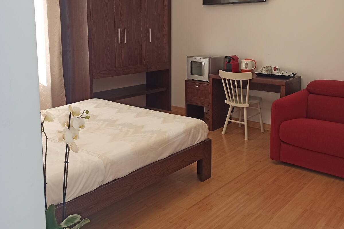 Ein Schlafzimmer mit einem Bett, einem Schreibtisch mit Fernseher, einem roten Sofa und einem Holzschrank.
