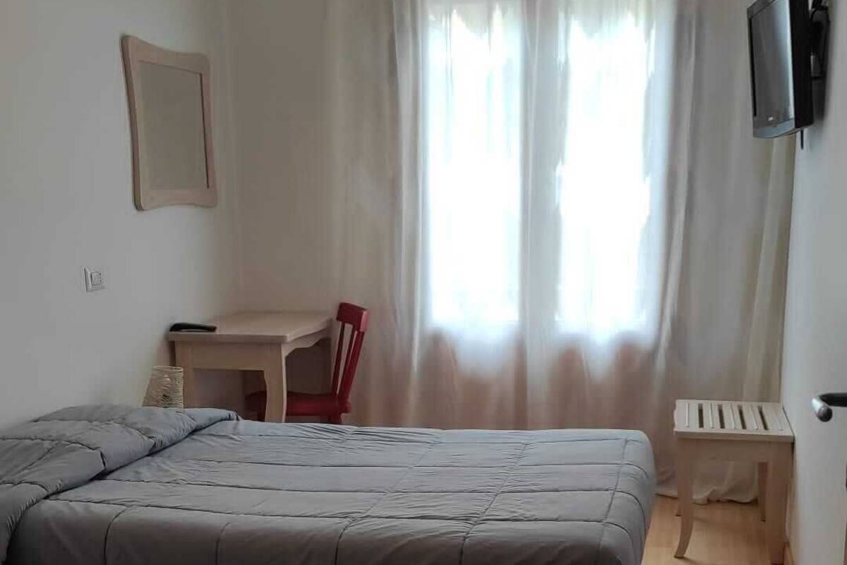 Ein Schlafzimmer mit Bett, Schreibtisch, Stuhl, Fernseher und einem Fenster mit Vorhängen.