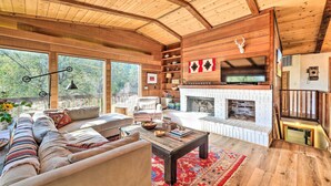 A spacious living room with a fireplace, wooden beams, and large windows. - Groveland Cabin w/ Outdoor Perks & Game Room (Groveland)