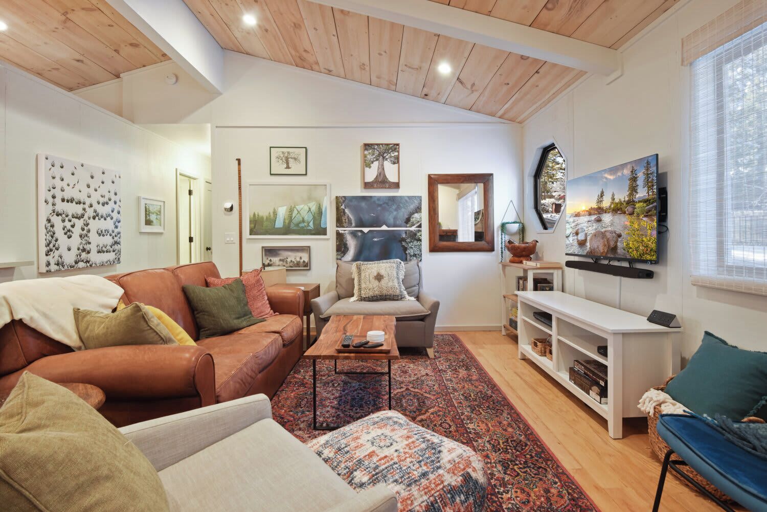 A cozy living room with a brown leather sofa, a grey armchair, and a patterned rug. There is a flat-screen TV mounted on the wall, a wooden coffee table, and a bookshelf with various items.