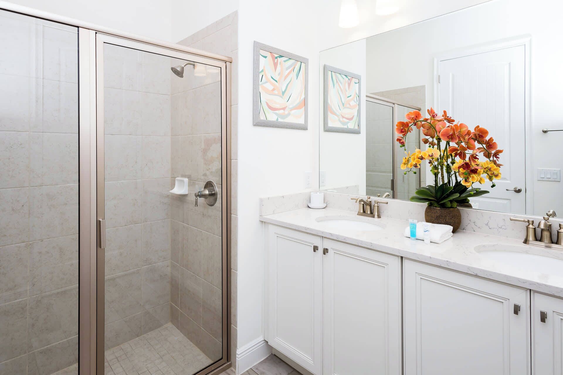Une salle de bain moderne avec une douche à parois de verre, des meubles blancs, un grand miroir et un vase rempli de fleurs.
