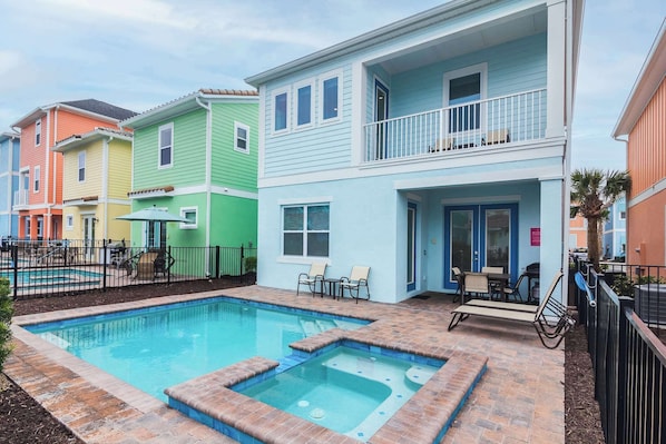 A pool area with a hot tub, outdoor seating, and colorful buildings in the background.