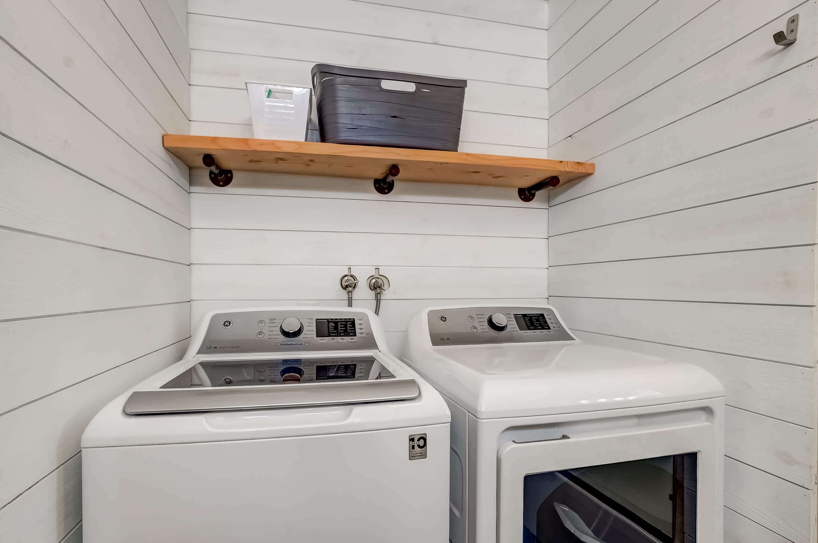 A laundry room with two washing machines, a wooden shelf with a basket, and a wall-mounted paper towel dispenser.