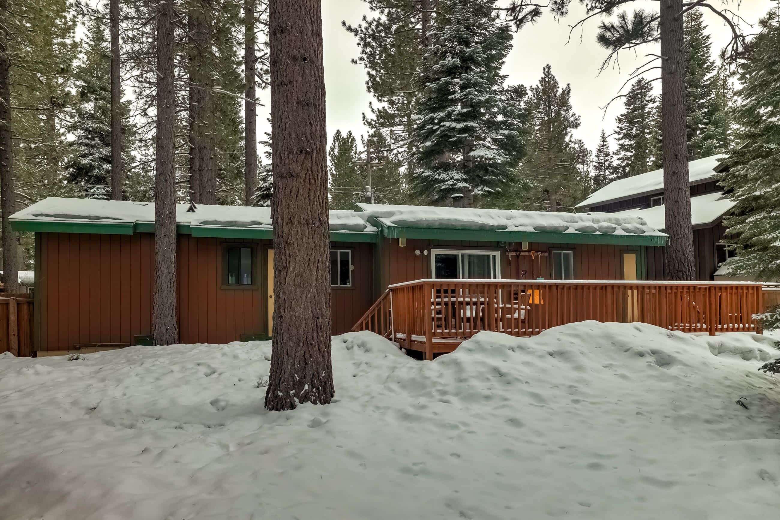 A cabin with a wooden deck and snow-covered roof, surrounded by tall trees.