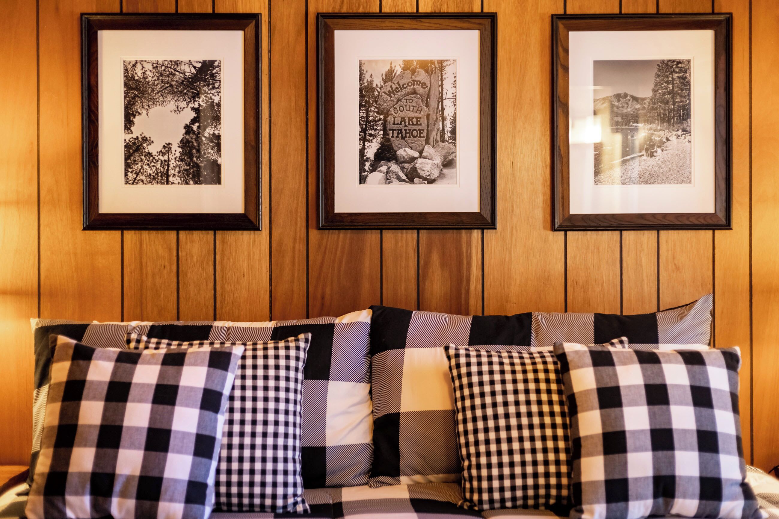A wooden paneled wall with three framed black and white photographs and a checkered sofa with multiple cushions.