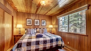 Un dormitorio con paneles de madera, colcha de cuadros, dos lámparas de noche, cuadros en la pared y un ventanal con vistas a un bosque.