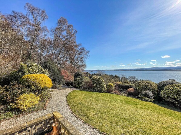 A landscaped garden with a pathway leading to a grassy area, surrounded by various shrubs and trees, overlooking a body of water.