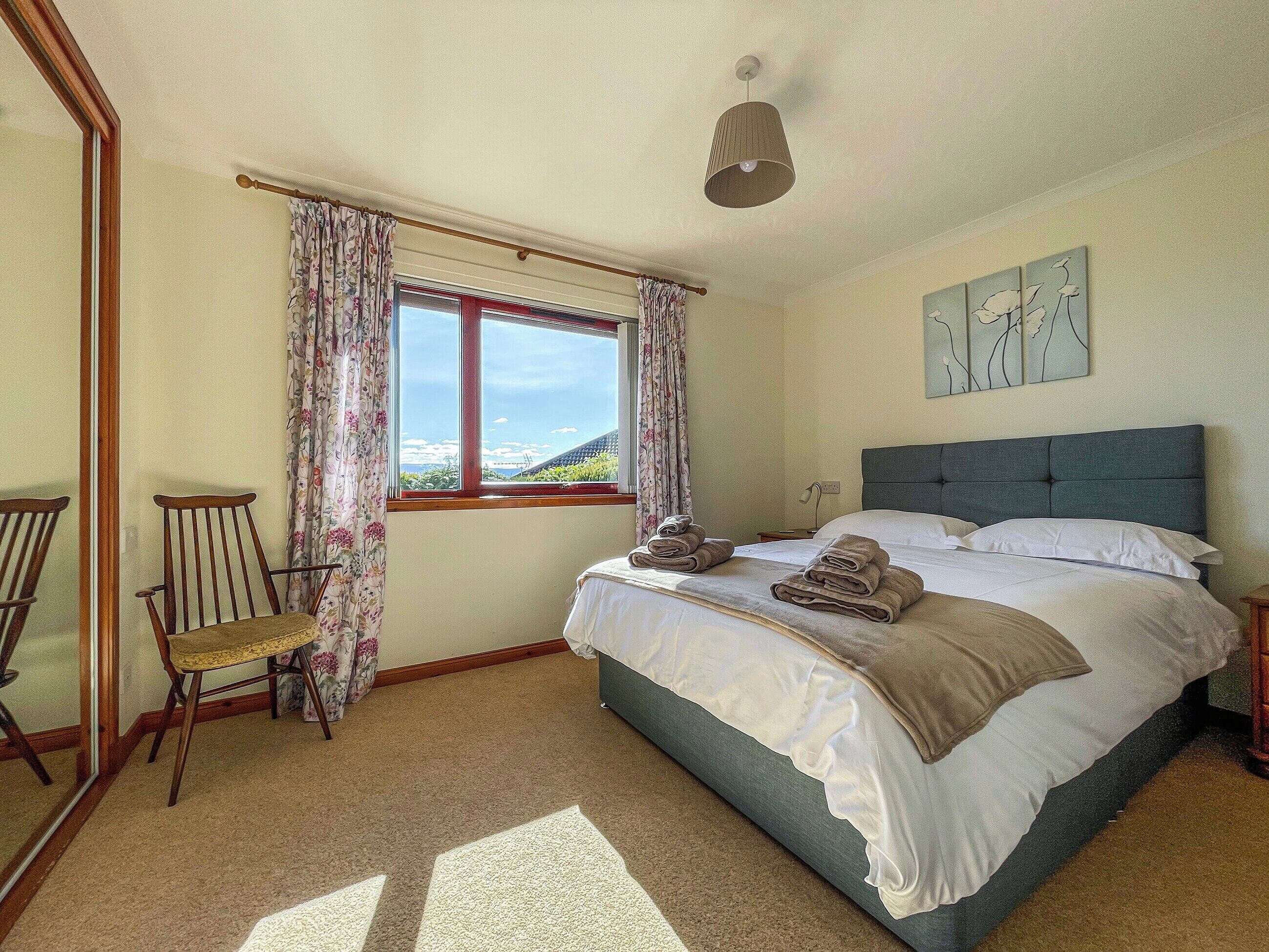 A bedroom with a bed, a chair, a window with curtains, and artwork on the wall.