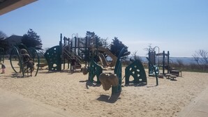 A playground with various equipment, including swings and climbing structures, set on sandy ground with a clear sky in the background.