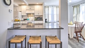 A modern kitchen with white cabinets, a granite countertop, and a built-in microwave. There are three woven stools at the counter and a dining area with a wooden table and chairs. - Peaceful Hurley Retreat ~ 1 Mi to Main Street (Hurley)
