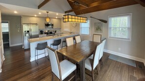 A spacious kitchen with a central island, wooden ceiling beams, and a dining area with a large wooden table and chairs. - The Cozy Cottage on Little Duck Cove (Roxboro)