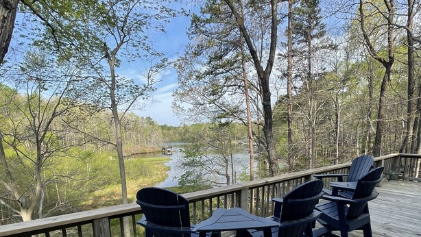 A wooden deck with four Adirondack chairs and a table, overlooking a calm lake surrounded by trees. - The Cozy Cottage on Little Duck Cove (Roxboro)