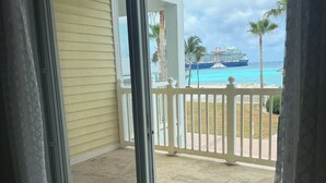 A view from a room with a sliding glass door to a balcony with a white fence, a pool, and a cruise ship in the background.