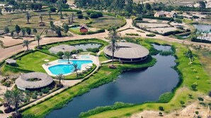 An aerial view of a resort with a central swimming pool, circular water features, and palm trees.