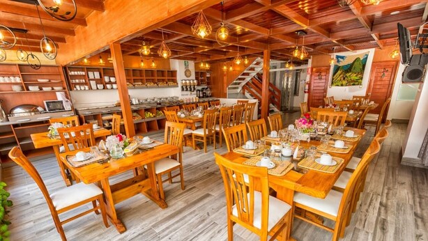 A spacious dining area with wooden tables and chairs, a kitchen in the background, and a staircase leading to an upper level. - Chakana Inn (Machu Picchu)