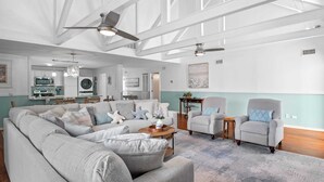 Ein geräumiges Wohnzimmer mit einem Ecksofa, Sesseln und einem Couchtisch. Es gibt einen Deckenventilator, Pendelleuchten und ein großes Fenster, das natürliches Licht hereinlässt.