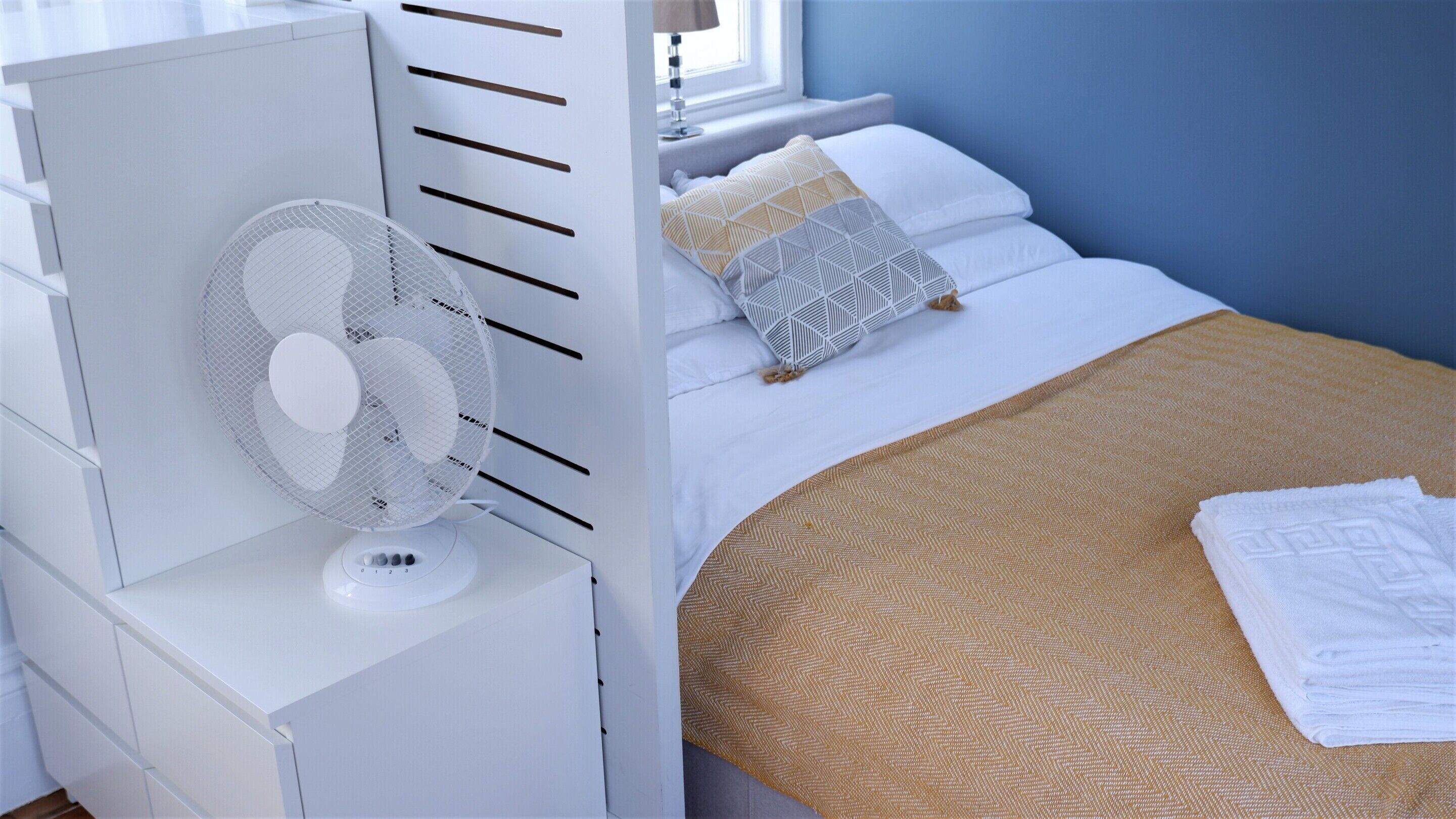 Un dormitorio ordenado con cama, almohadas, una mesita de noche blanca y un ventilador de pie.