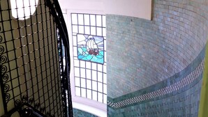 Una escalera con barandilla de hierro forjado negro, un muro con mosaicos y un ventanal de vidrio emplomado.