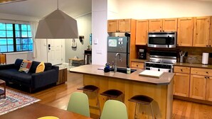 Eine moderne Küche mit Holzschränken, einem Edelstahlkühlschrank, einer Mikrowelle und einem Gasherd. Es gibt eine Essecke mit Tisch und Stühlen sowie ein Wohnzimmer mit Sofa und Fenster.