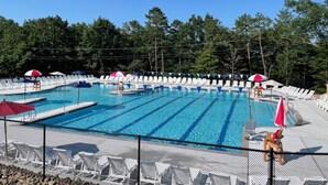Ein Schwimmbecken mit mehreren Bahnen, umgeben von Liegestühlen und Sonnenschirmen. Im Hintergrund ist ein Zaun und Bäume zu sehen.