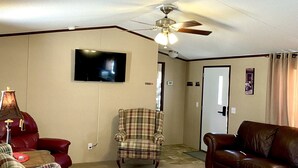 A living room with a ceiling fan, a television, a plaid armchair, a red leather chair, and a brown leather couch. - Just Dune It - East Home (Waynoka)