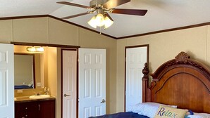 A bedroom with a wooden bed, a ceiling fan, and a bathroom visible through an open door. - Just Dune It - East Home (Waynoka)