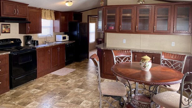 A compact kitchen with dark wooden cabinets, a black refrigerator, a microwave, and a stove. There is a dining table with four chairs and a decorative vase. - Just Dune It - East Home (Waynoka)