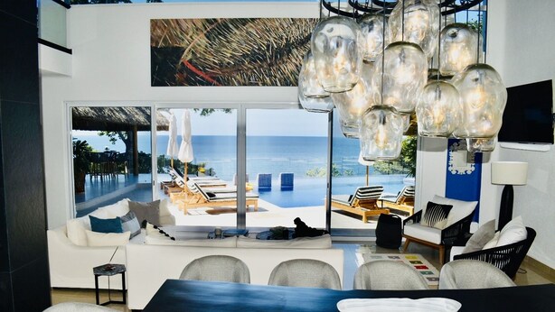 A modern living room with a large window overlooking a pool and ocean view. There are multiple seating areas, a dining table, and a hanging light fixture. - Exclusive Beach Front Luxe Boutique Retreat (Bahia de Banderas)