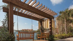 Een houten pergola met schommels kijkt uit over een waterpartij, omgeven door groen en hoge gebouwen op de achtergrond.