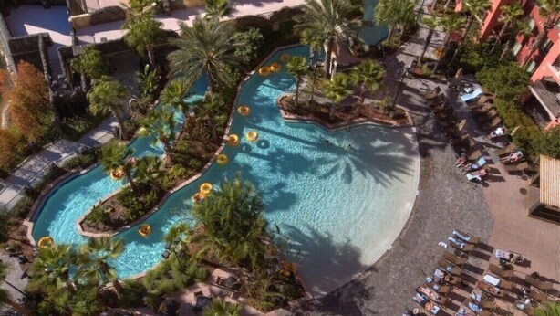Luchtbeeld van een resort met een kronkelend zwembad, omgeven door palmbomen en terrasjes.