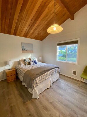 A bedroom with a wooden ceiling, a bed with a checkered bedspread, a bedside table with a lamp, a window with a view of greenery, and a green chair.